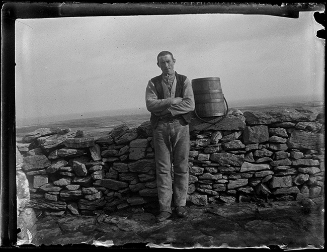 John Millington Synge took this  photograph of a young man a posing alongside a drystone wall that is traditional in the Aran Islands. He is dressed in traditional home spun vest, shirt, waist coat and trousers. The man wears rawhide sandals while the boy wears boots. Lilo Stephens identified the man as Martin McDonagh. Photo Credit:John Millington Synge. 1898, Digital photographs from scanned silver gelatine negatives (Timothy Keefe, Sharon Sutton 2009). Courtesy of the Board of Trinity College, University of Dublin. Alt text by Ciarán Walsh, curator.ie.