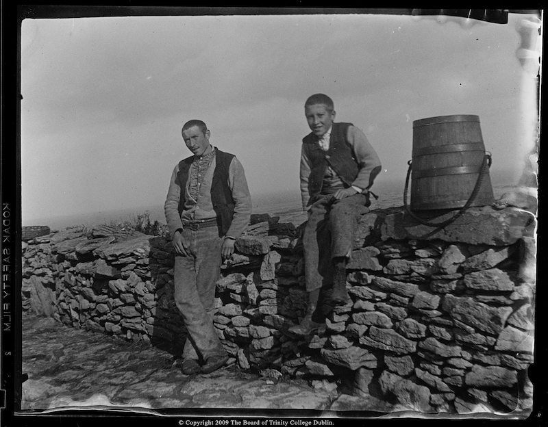 John Millington Synge took this  photograph of a young man and a boy posing alongside a drystone wall that is traditional in the Aran Islands. Both are dressed in traditional home spun vest, shirt, waist coat and trousers. The man wears rawhide sandals while the boy wears boots. Lilo Stephens identified the man as Martin McDonagh. photo credit: John Millington Synge. 1898, Digital photographs from scanned silver gelatine negatives (Timothy Keefe, Sharon Sutton 2009). Courtesy of the Board of Trinity College, University of Dublin. Alt text by Ciarán Walsh, curator.ie.